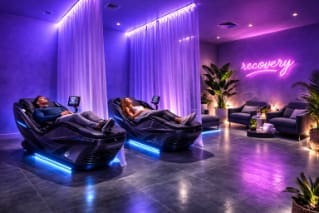 Purple‑lit recovery room with two people on high‑tech massage loungers.